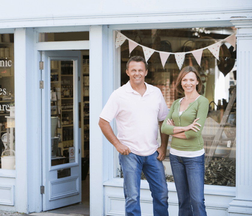 Business-Owners Smiling man and woman standing outside their small business storefront with light blue trim and decorative bunting in the window.