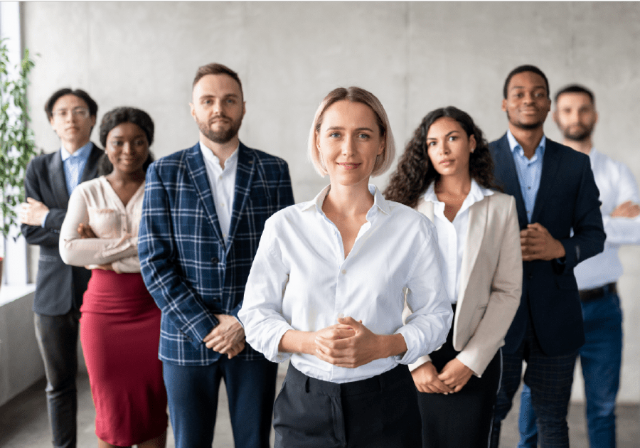 Diverse group of confident business professionals standing together in formal attire, with a woman in a white blouse leading in front.