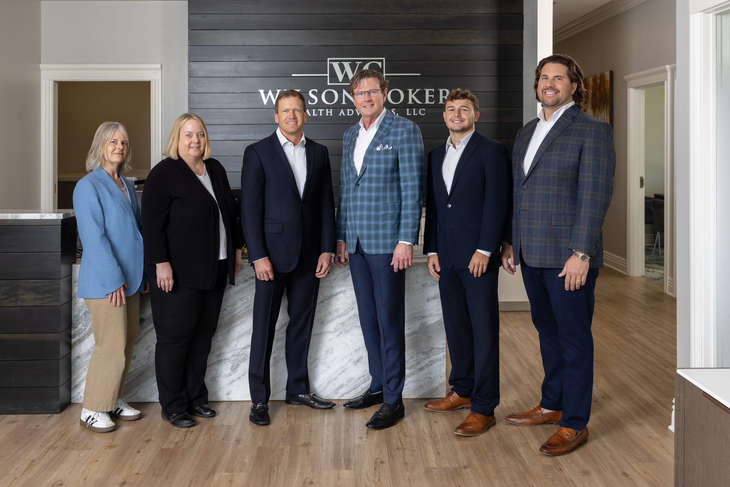 Six professionals in business attire pose for a group photo in a modern office lobby with a dark wood wall behind them and a sign.